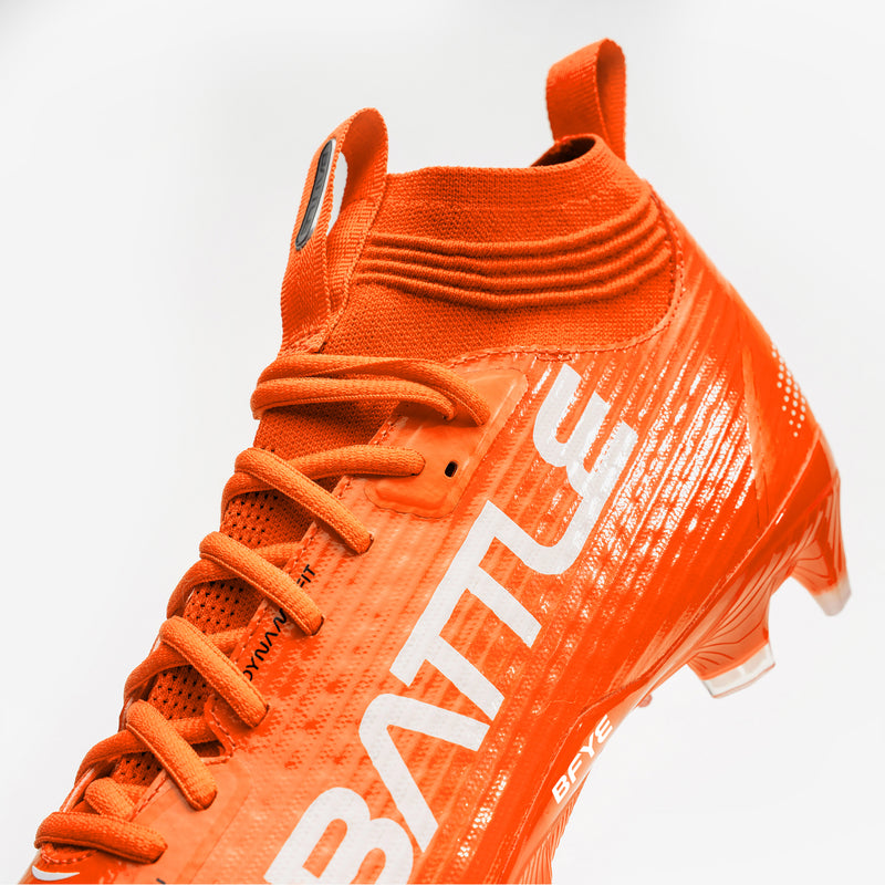 A pair of bright orange football cleats with white 'BATTLE', displayed on a clean white background.